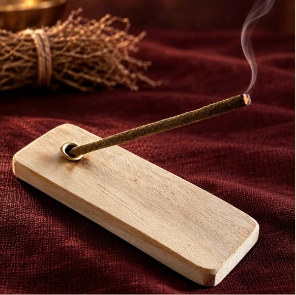 A lit Udupi Sugandha incense stick resting at an angle in its wooden holder on maroon cloth, beside a bundle of lavancha roots